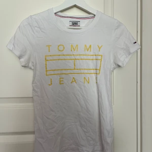 Vit t-shirt Tommy Jeans XS - Vit t-shirt från Tommy Jeans i storlek XS med gul logotypprint framtill. Klassisk rund hals och korta ärmar. Liten Tommy Jeans-flagga på ärmen. Mjuk bomullskvalitet som är skön mot huden. Perfekt för en clean och fräsch look.
