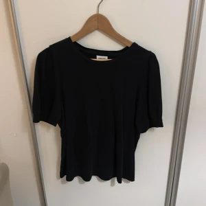 Svart blus från AWARE med puffärm - Svart blus från AWARE med rund hals och korta puffärmar. Stilren och enkel design med mjukt, slätt tyg som faller snyggt. Perfekt för dig som gillar minimalistisk stil och vill ha en topp som är lätt att matcha med olika outfits.