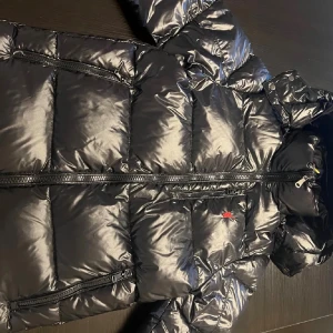 Svart pufferjacka Polo Ralph Lauren XS - Svart pufferjacka från Polo Ralph Lauren i storlek XS. Jackan har glansig yta, huva, flera dragkedjefickor och klassisk röd Polo-logga på bröstet. Materialet är syntet och dun, perfekt för kalla dagar. Snygg och stilren modell med lång ärm och dragkedja framtill.