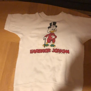 Vit Farbror Joakim tröja Disney - Vit kortärmad tröja med tryck av Farbror Joakim och texten 'Farbror Joakim' i rött framtill. Tröjan har rund hals och är gjord i mjuk bomull, perfekt för dig som gillar retro och Disney-vibes.