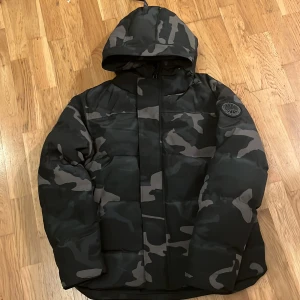 Camouflage dunjacka från Canada Goose - Säljer en fet dunjacka från Canada Goose med mörkt camouflage-mönster och huva. Jackan har en snygg patch på ärmen och dragkedja framtill. Perfekt för kalla dagar och riktigt street. Kommer med originalpåse och tag.