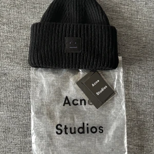 Acne studios mössa - Svart ribbstickad mössa från Acne Studios med ikonisk ansiktslogga framtill. Tillverkad i 100% ull för extra värme och stil. Perfekt accessoar för att lyfta din look under kyliga dagar. Kommer med originalpåse och tag.