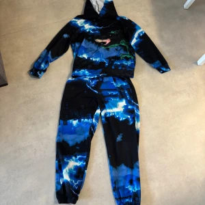 Blå hoodie med blixtmotiv från SHEIN - Kolla gärna in mina andra atiklkar! Säljer en cool hoodie från SHEIN i storlek 11Y med ett fett blått och svart blixtmönster och en stor grön dinosaurie på framsidan. Tröjan har huva och långa ärmar, perfekt för dig som gillar unika prints och streetstyle.