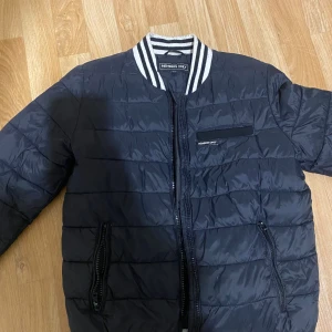 Svart quiltad bomberjacka Members Only - Svart quiltad bomberjacka från Members Only med vita och svarta ränder på kragen. Jackan har dragkedja framtill, två sidofickor med dragkedja och en bröstficka med logga. Tillverkad i syntetmaterial och har långärmad design.