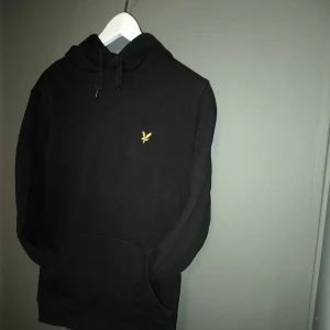 Svart hoodie från Lyle & Scott - Svart hoodie från Lyle & Scott med klassisk gul logga på bröstet. Tröjan har huva med snörning och en stor magficka framtill. Tillverkad i mjuk bomull och har långa ärmar. Perfekt för en chill och stilren look.