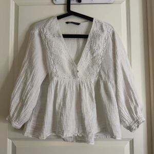 Vit blus med spets från Zara - Säljer en vit blus från Zara med v-ringning och broderade spetsdetaljer framtill. Blusen har långärmade puffärmar med resår vid ärmslut och en luftig, flowy passform. Materialet är mjukt och lätt, perfekt för vår och sommar.