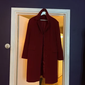 Vinröd kappa i ullmix, strl 46 - Snygg vinröd kappa med klassisk krage och knäppning framtill. Jackan har två fickor med lock och är tillverkad i en mjuk ullmix med polyamid. Fodret är i acetat. Perfekt för dig som vill ha en stilren och tidlös look.