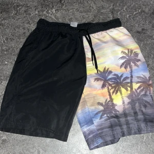 Svarta och mönstrade badshorts/shorts med snörning - Säljer ett par unika badshorts med ena benet helsvart och andra benet med färgglatt palmtryck i solnedgångstoner. Shortsen har elastisk midja och svart snörning för perfekt passform. Perfekta för stranden eller poolen!