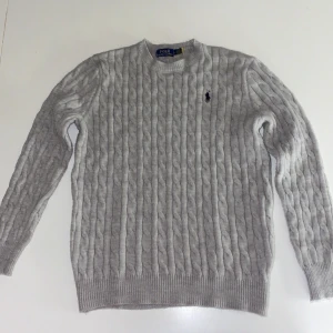 Polo Stickad tröja grå - Snygg grå kabelstickad tröja från Polo Ralph Lauren i storlek L. Tröjan har rund hals, lång ärm och klassisk broderad logga på bröstet. Riktigt bra kvalité 