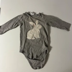 Grå body med kaninapplikation - Supersöt grå body med lång ärm och tryckknappar i grenen. Framsidan har en stor vit kaninapplikation med en liten rosa rosett vid örat. Perfekt för dig som gillar gulliga detaljer och mjuka material.