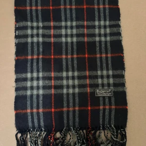 Rutig ullhalsduk från Burberry - Snygg rutig halsduk från Burberry i 100% lammull. Mörkblå bas med grå och röda rutor, klassisk design och fransar i ändarna. Perfekt accessoar för att lyfta din stil under kyliga dagar. halsduken skickad från Norge och moms kan tilkomma. betalning måste ske via paypal eller liknande