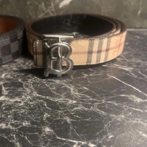 Burberry rutigt bälte med silverdetalj - Snyggt bälte från Burberry med klassiskt beige, brunt och rött rutmönster samt en stor silvrig spänne med TB-logga. Perfekt accessoar för att lyfta din outfit med en lyxig touch. Bältet är gjort i skinn och har en stilren design.