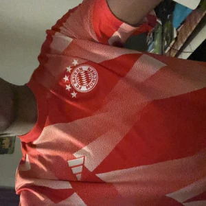 Röd FC Bayern München fotbollströja - Säljer en snygg FC Bayern München fotbollströja från Adidas. Tröjan är röd med vita geometriska mönster och har klubbmärket med fyra stjärnor på bröstet. Kortärmad modell i lätt och ventilerande material, perfekt för träning eller match.