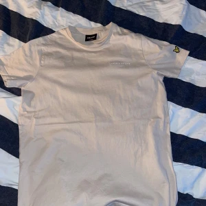 Beige t-shirt från Lyle & Scott - Snygg beige t-shirt från Lyle & Scott med broderad logga på bröstet och den klassiska gula fågeln på ärmen. T-shirten har rund hals och är kortärmad, tillverkad i mjuk bomull för skön känsla. Perfekt för en clean och stilren look.