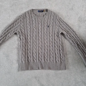 Polo Ralph Lauren - Kabelstickad beige tröja från Polo Ralph Lauren med rund hals och långärmad passform. Tröjan har det klassiska broderade Polo-logot på bröstet och ribbade muddar vid ärmslut och nederkant. Perfekt för dig som gillar stilren och tidlös design. Storlek S personen på bilden är 181