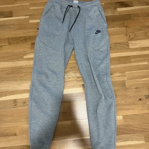 Grå mjukisbyxor från Nike tech - Grå mjukisbyxor från Nike tech i storlek S med svart Nike-logga på vänster ben. Byxorna har elastisk midja med svart snörning, två sidofickor och är tillverkade i mjukt bomullsmaterial. Perfekta för chill eller träning. Knappt använda och inga defekter eller sådant!