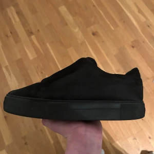 Svarta slip-on sneakers från Arigato - Säljer ett par svarta slip-on sneakers från Arigato med platt sula och rund tå. Skorna har en stilren design utan snörning och är tillverkade i mocka med elastiska paneler för enkel påtagning. Perfekt för dig som gillar minimalistisk och clean stil.