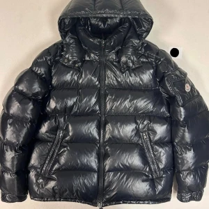 Moncler Maya - Svart pufferjacka från Moncler, modell Maya. Jackan har en glansig finish, huva, dragkedja framtill och två snedställda fickor med dragkedja. Moncler-logga på ärmen och ett unikt serietidningsmärke på insidan. Perfekt för kalla dagar med stil. Självklart äkta storlek 6/7 då lappen har gått av  ligger på 18 tusen nya 