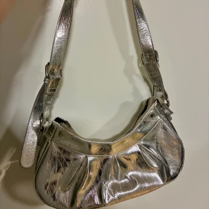 Silvrig axelväska från Gina Tricot -  Handväska i fint skick i metallic-silver från Gina Tricot. Väskan har en justerbar axelrem, dragkedja upptill och snygga veckade detaljer på framsidan. 