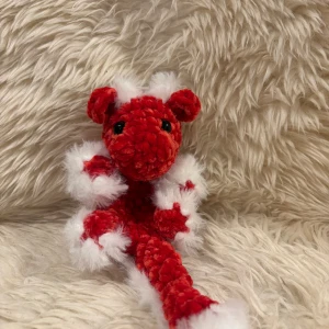 Virkad mini dragon snuggler  - Bilderna visar ett rött och vitt virkat gosedjur, drake eller fantasidjur, med svarta ögon och fluffiga detaljer. Produkten ser handgjord ut och är i mycket gott skick utan synligt slitage. Perfekt som present eller dekoration.