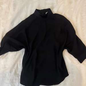 Svart blus från Carin Wester - Säljer en svart blus från Carin Wester i storlek 38. Toppen har en krage och är tillverkad i ett mjukt, slätt material. Modellen är loose fit med trekvartsärmar och dragkedja i nacken. Perfekt för dig som gillar minimalistisk och stilren design.