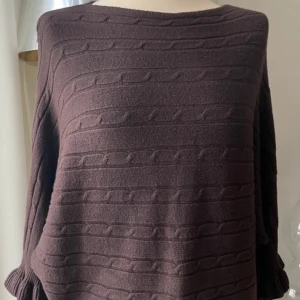 Brun stickad tröja med volangärm - Mysig brun stickad tröja med snyggt kabelmönster och unika volangdetaljer vid ärmsluten. Tröjan har en loose passform och rund halsringning, perfekt för dig som gillar en chill och trendig look.