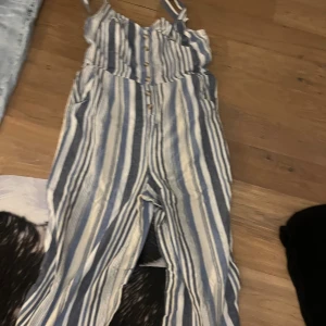 Randig jumpsuit med knappar - Snygg jumpsuit med blå och vita ränder, knappar framtill och smala axelband. Lätt och luftig modell som passar perfekt till varma dagar. Raka ben och fickor i sidorna ger en avslappnad vibe.