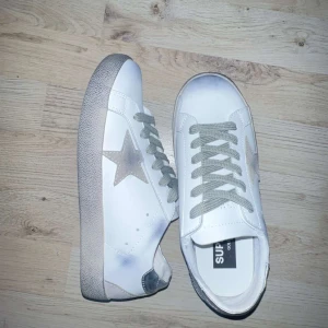 Vita sneakers med stjärndetalj - Snygga vita sneakers med grå snörning och en grå stjärna på sidan. Skorna har en platt sula och är tillverkade i syntetmaterial med en cool, lite sliten look. Perfekta för dig som gillar streetstyle och vill sticka ut med en unik detalj.