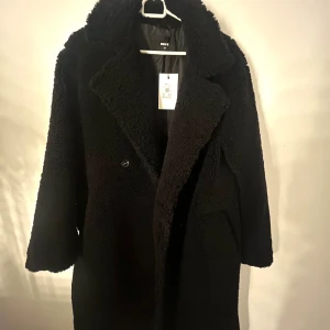 Svart teddyjacka från Bik Bok - Säljer en svart teddyjacka från Bik Bok i oversized modell. Jackan har bred krage, två knappar framtill och stora fickor. Materialet är mjukt och fluffigt, perfekt för kalla dagar. Passformen är loose och ärmarna är långa. Riktigt snygg och trendig jacka för dig som gillar streetstyle.