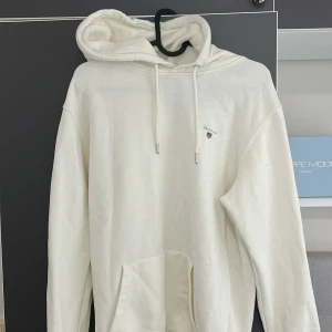 Vit hoodie från GANT med huva - Säljer en stilren vit hoodie från GANT med broderad logga på bröstet. Tröjan har stor huva med snörning, magficka och ribbade muddar vid ärmslut och nederkant. Perfekt för chill dagar och enkel att matcha med jeans eller joggers. Den är knappt använd kanske 5 gånger max och är i perfekt skick inga defekter eller fläckar nån stans. Pris är inte hugget än, billigare vid snabb affär. Hör av dig vid funderingar.🤩