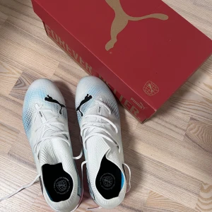 Puma vita fotbollsskor med rosa sula - Säljer ett par snygga vita fotbollsskor från Puma med svart logga och coola blå detaljer. Skorna har snörning och en platt, rosa sula med dobbar för grymt grepp på planen. Materialet är syntet och designen är modern med grafiska inslag på sidan.