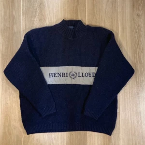 Mörkblå stickad tröja Henri Lloyd - Snygg mörkblå stickad tröja från Henri Lloyd med bred beige panel och logga över bröstet. Tröjan har ribbad hals, muddar och nederkant samt långa ärmar. Perfekt för dig som gillar klassisk stil med en twist.