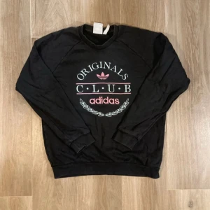 Svart sweatshirt från Adidas Originals - Svart sweatshirt från Adidas Originals med tryck i rosa och vitt framtill. Tröjan har rund hals, långa ärmar och ribbade muddar vid ärmslut och nederkant. Perfekt för en chill och sportig stil.