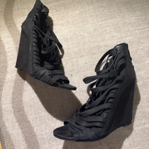 Svarta wedge sandaler med snörning - Säljer ett par svarta sandaler med höga wedge-klackar och snygga snörningsdetaljer framtill. Skorna har öppen tå och flera breda remmar i mocka som ger en cool och edgy look. Perfekta för dig som vill sticka ut med din stil.
