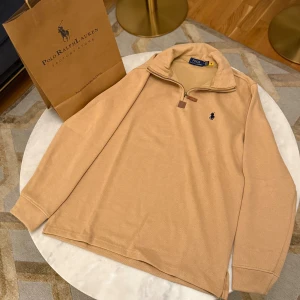 Beige half zip tröja Polo Ralph Lauren - Snygg beige långärmad tröja från Polo Ralph Lauren med klassisk broderad logga på bröstet. Tröjan har en half zip-dragkedja vid kragen och är gjord i mjuk bomull. Perfekt för dig som gillar stilrena och tidlösa plagg.