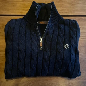 Svart kabelstickad Morris halfzip - Säljer en Svart halfzip tröja från Morris. Tröjan har ett klassiskt kabelstickat mönster och en broderad logotyp på bröstet. Perfekt för kyliga sommar kvälller, men funkar även till svala höst och vår dagar. 
