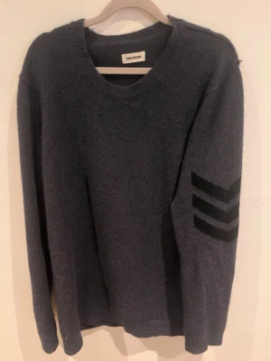 Mörkgrå stickad tröja från Zadiq voltiere - Mörkgrå stickad tröja från Zadiq voltiere med rund hals och lång ärm. På ena ärmen finns svarta chevron-detaljer som ger en cool touch. Perfekt för dig som gillar enkel men snygg stil. Mjuk och skön kvalitet. Oversized