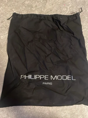 Svart dustbag från Philippe Model - Säljer en svart dustbag från Philippe Model Paris. Väskan har en enkel design med dragsko upptill och logotypen broderad i vitt på framsidan. Perfekt för att förvara och skydda dina skor.