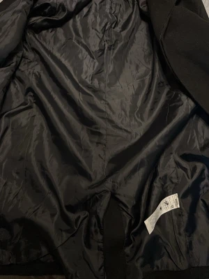 Svart kappa från Pier One - Svart kappa från Pier One med klassisk rak passform och knappar framtill. Yttermaterialet är mjukt och känns lyxigt, fodret är slätt och skönt mot kroppen. Perfekt för dig som vill ha en stilren och enkel look under höst och vinter.