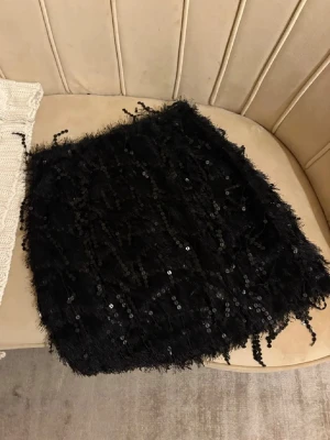 Svart paljett kjol XS - Säljer en svart palljet kjol i storlek XS med glittriga paljetter och fransiga detaljer. Kjolen en har en cool textur och är axelbandslös, perfekt för att sticka ut.  Helt ny använde bara på nyår