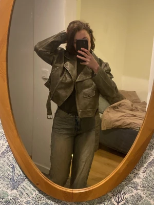 Olivgrön, med bruna toner skinnjacka från Urban Classics - Säljer en olivgrön bikerinspirerad skinnjacka från Urban Classics i storlek M. Jackan har klassisk krage, dragkedjor och ett bälte i midjan. Snygg croppad passform loose fit/avslappnad sitter inte tajt på kroppe. Som ger en cool vibe till din outfit.