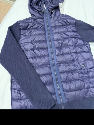 Moncler cardigan - Säljer en mörkblå pufferjacka från moncler med stickade ärmar och huva. Jackan har dragkedja framtill, ribbade muddar och en extra ficka med dragkedja på ena ärmen. .