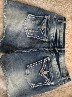 Blå denimshorts med slitningar - Säljer ett par blå denimshorts med fransiga kanter och slitna detaljer. Shortsen har stora fickor med dekorativa sömmar och silverfärgade knappar. Passar dig som gillar en avslappnad och cool stil till sommaren.