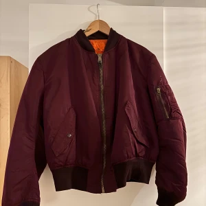 Vinröd bomberjacka med dragkedja - Säljer en mörk vinröd bomberjacka med klassisk orange insida, ribbade muddar och dragkedja framtill. Jackan har fickor med tryckknappar och en extra ficka med dragkedja på ärmen. Perfekt för dig som vill ha en cool och tidlös look.
