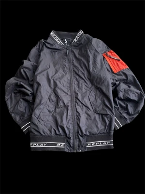 Svart jacka från Replay - Svart bomberjacka från Replay med coolt rött fack på ärmen och Replay-logga på krage och muddar. Jackan har dragkedja framtill, två fickor med knapp och är tillverkad i lätt polyester. Perfekt för dig som vill ha en streetig look. Passar 158 cm 14y