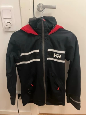 Svart och röd vindjacka från Helly Hansen - Säljer en svart vindjacka från Helly Hansen med röda detaljer och huva. Jackan har vita reflexränder över bröstet och är utrustad med dragkedja framtill samt praktiska fickor. Perfekt för blåsiga dagar och har en sportig look med HH-logga både fram och bak.