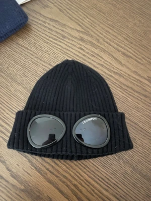 Svart ribbstickad mössa C.P. Company - Svart ribbstickad mössa från C.P. Company med ikoniska glasögon-detaljer framtill. Mössan är tillverkad i mjuk ull och har en uppvikt kant. Perfekt för dig som vill ha en unik och trendig accessoar med streetwear-vibe. OBS! DU VET VARFÖR DEN ÄR BILLIG!