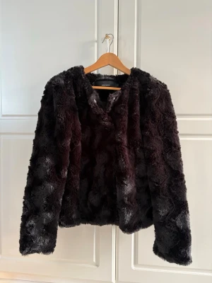 Svart pälsjacka från Vero Moda L - Mysig svart pälsjacka från Vero Moda med fluffig känsla och rund halsringning. Jackan har långa ärmar och stängs med tryckknappar framtill. Perfekt för dig som vill ha en stilren och trendig look under kyliga dagar.