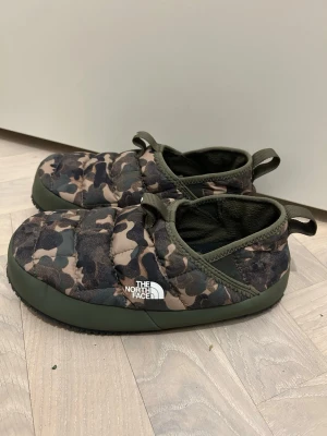 Camouflage slip-ons från The North Face 36 - Säljer ett par varma och sköna quiltade slip-on tofflor från The North Face med camouflage-mönster i gröna och bruna toner. Skorna har platt sula i svart gummi och praktiska dragflikar både fram och bak. Utsidan är i syntetmaterial och insidan är mjukt fodrad. Som nya, jättebra skick. Storlek 36.