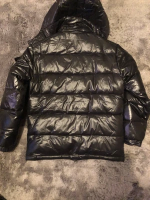 Svart Moncler pufferjacka med huva - Säljer en svart pufferjacka från Moncler med glansig finish och huva. Jackan har dragkedja framtill, långa ärmar och Moncler-logga på vänster arm. Perfekt för kalla dagar och har en riktigt schysst streetstil/grishstil